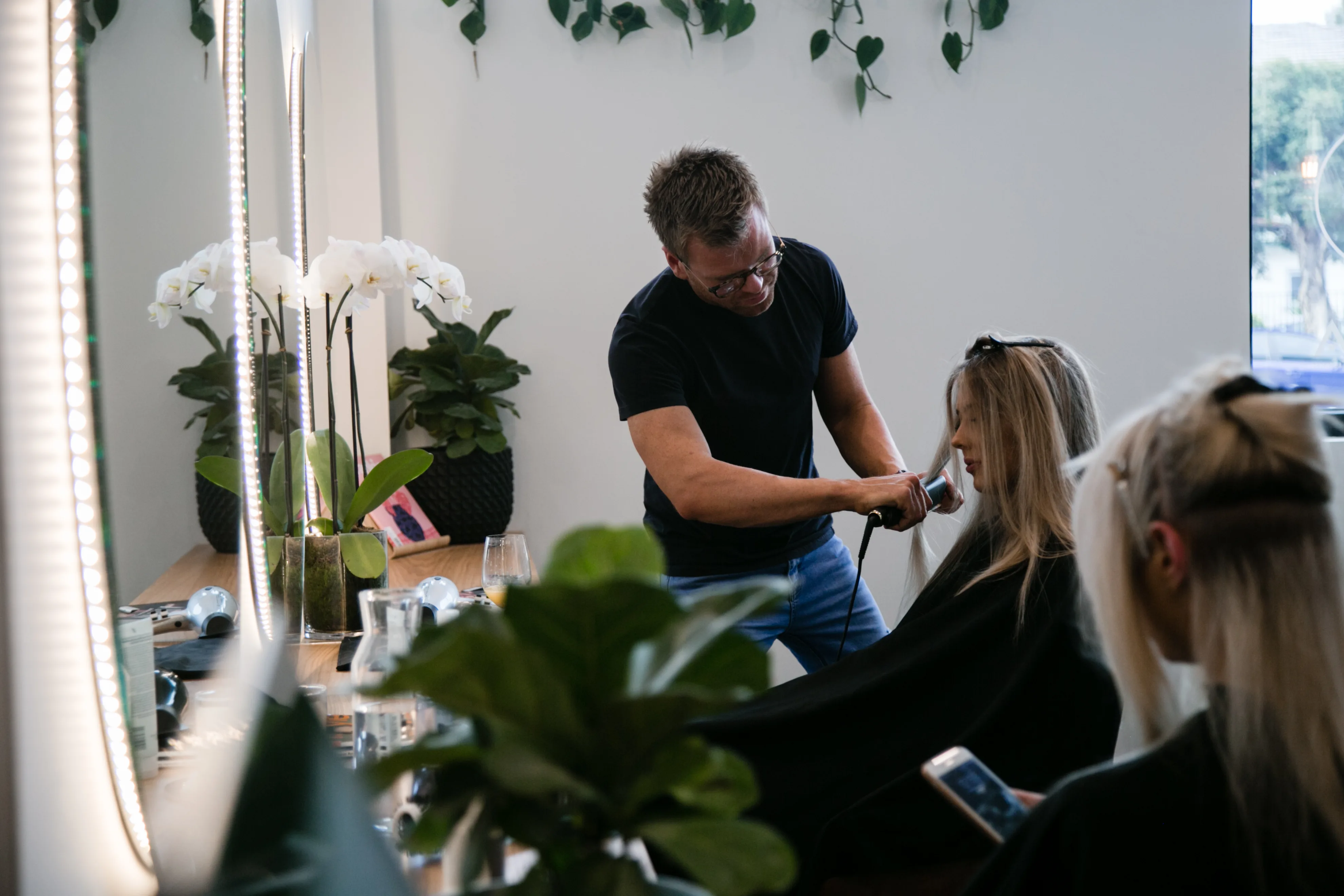 Stylist flat-ironing a blonde client with white orchids in background