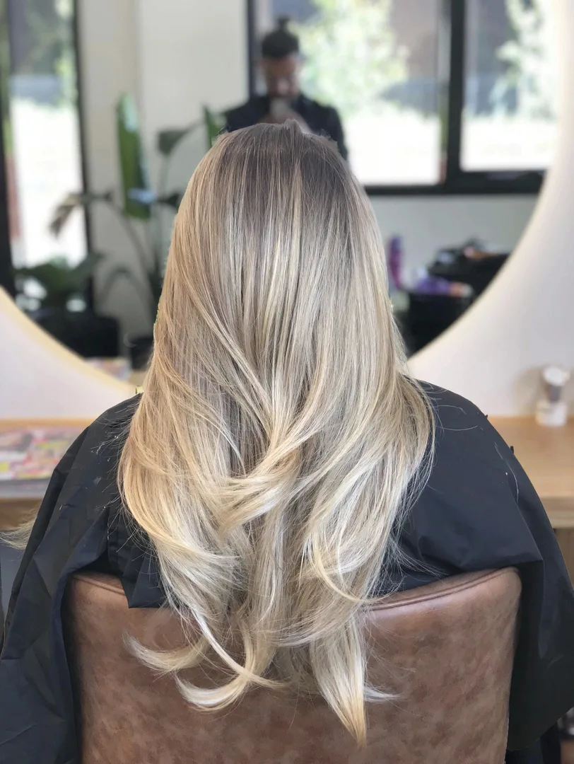 Long blonde balayage result in the styling chair, stylist reflected in round mirror